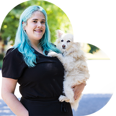 A person with long blue hair, dressed in a black outfit, holds a fluffy cream-colored dog. Likely a veterinarian, they stand outside against a blurred backdrop of trees and buildings.