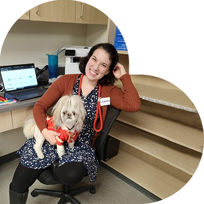 A woman sitting in an office chair holds a small dog wearing a red outfit on her lap. She is smiling, and a laptop is open on the desk beside her. There are shelves behind her with several boxes on them.